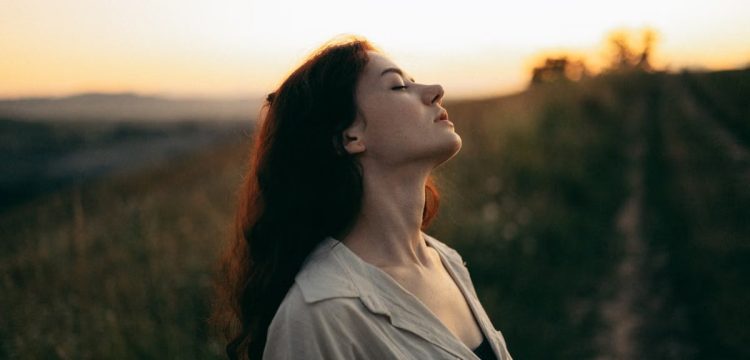 Woman standing with eyes closed in a field during sunset, exuding tranquility and peace.